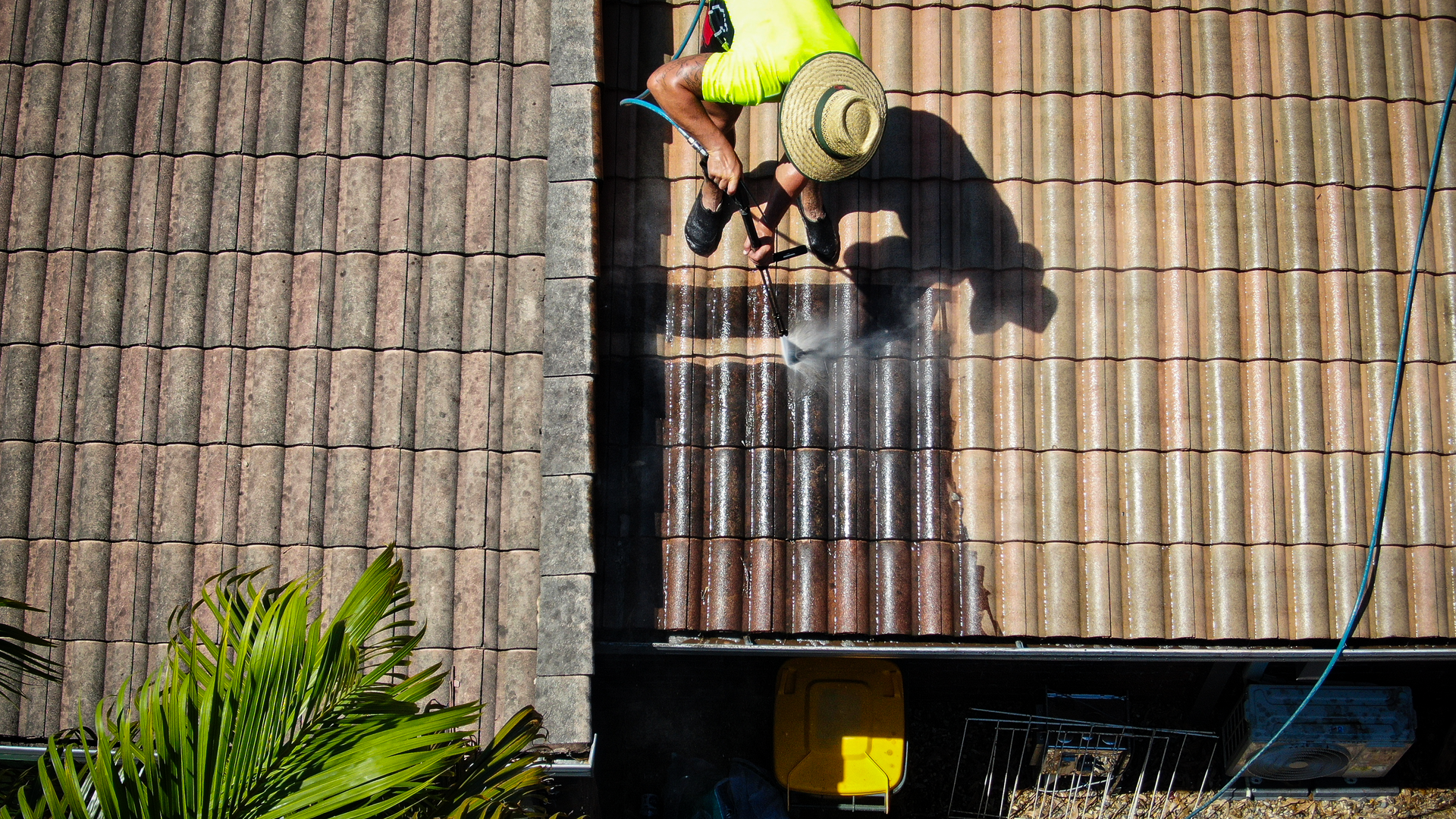 Worker on a roof pressure-washing shingles beside a palm tree