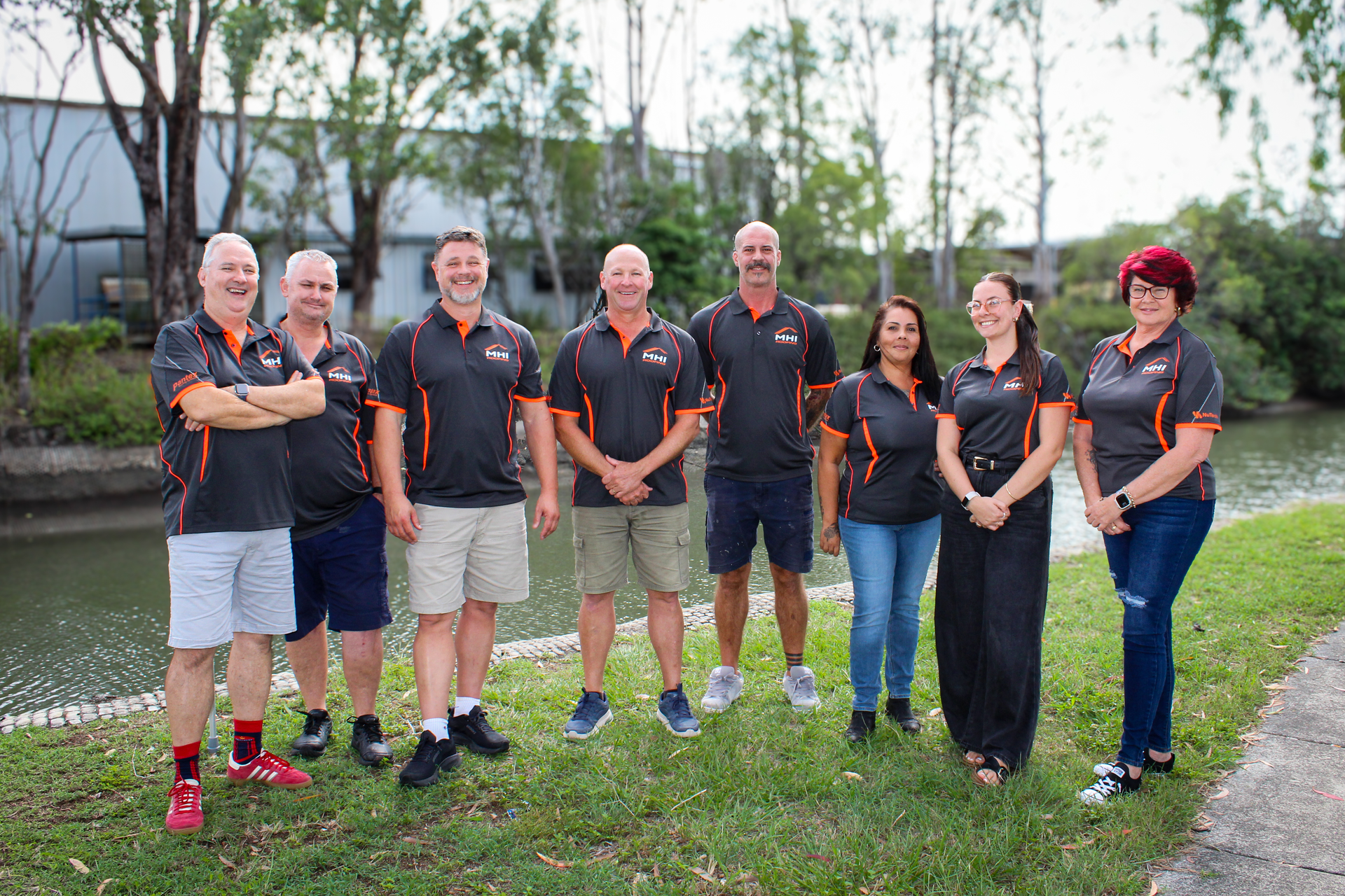 Group of seven people in matching black shirts standing outdoors by a pond and trees