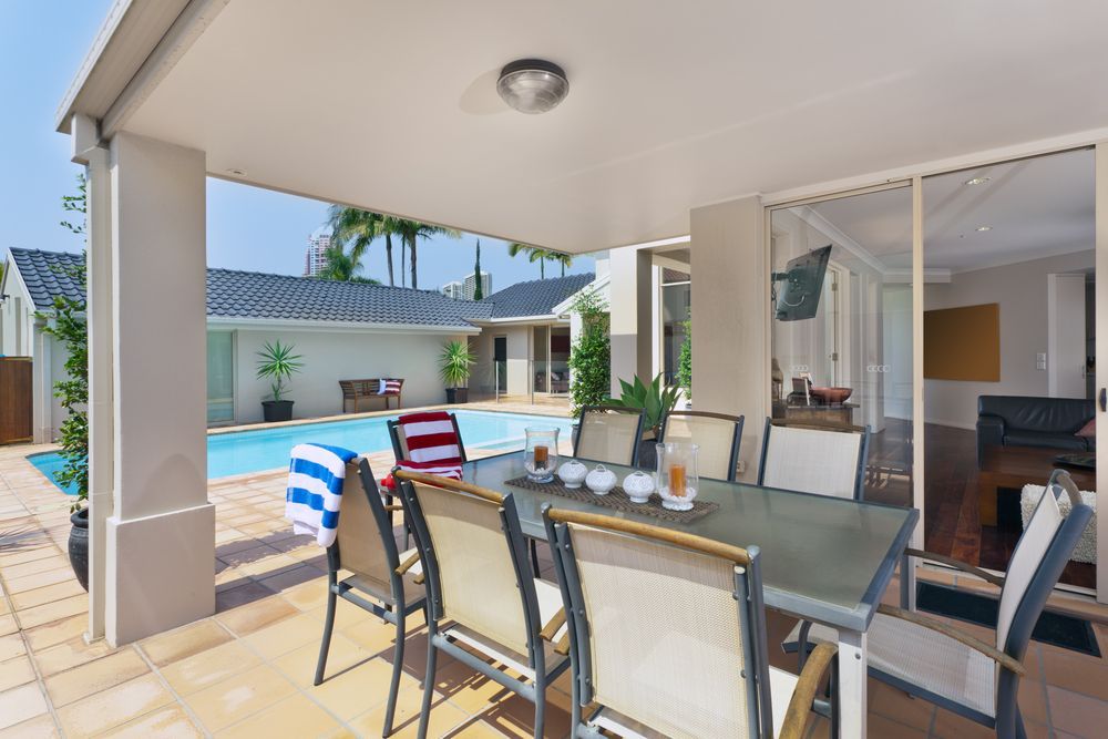 Covered patio with glass dining table, cushioned chairs, and poolside view in a sunny backyard