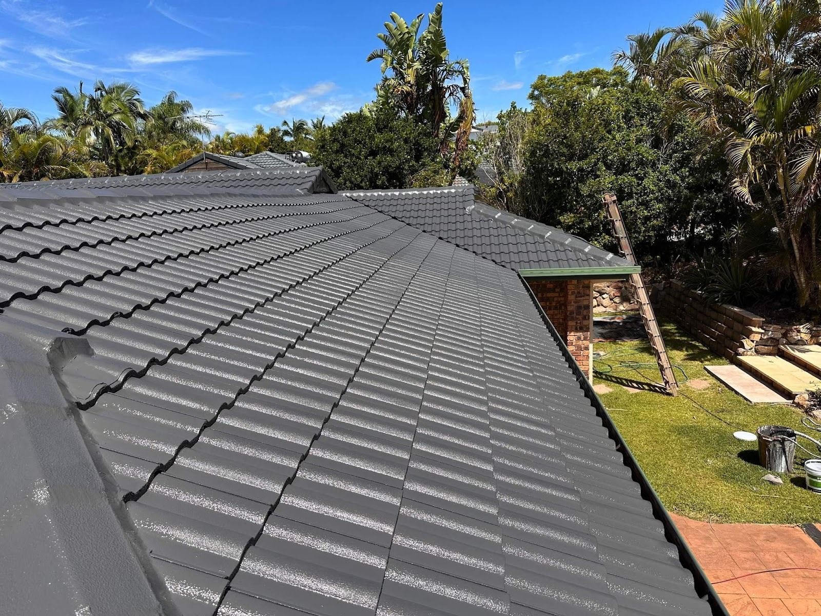 Dark gray tiled roof with solar panels beside a backyard and trees under a sunny sky
