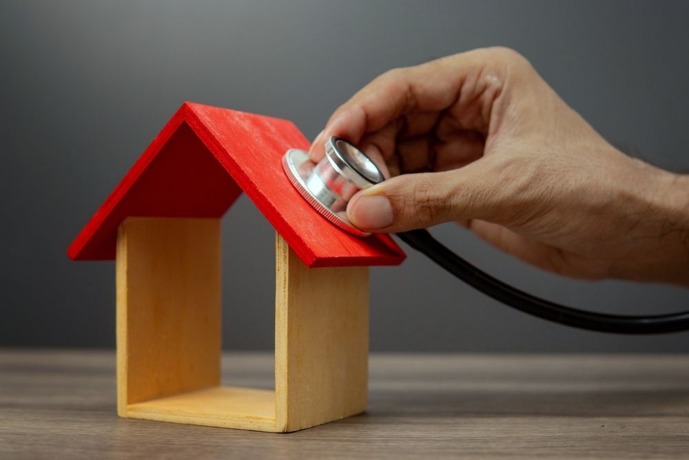 Hand holding a stethoscope to a small wooden house with a red roof on a gray surface