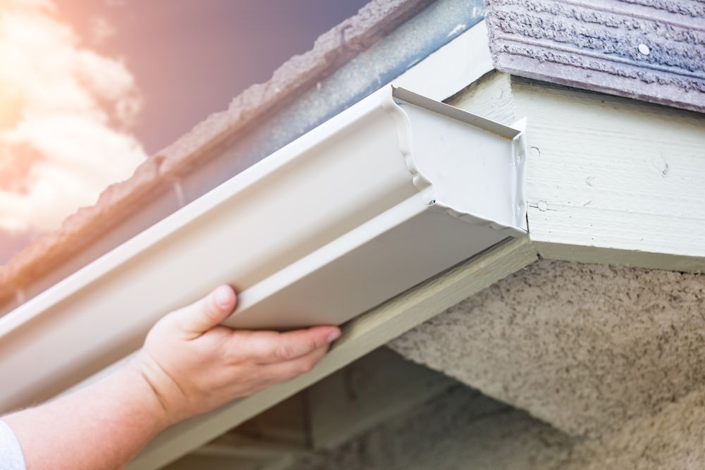Hand holding a white gutter section under a roof eave during installation