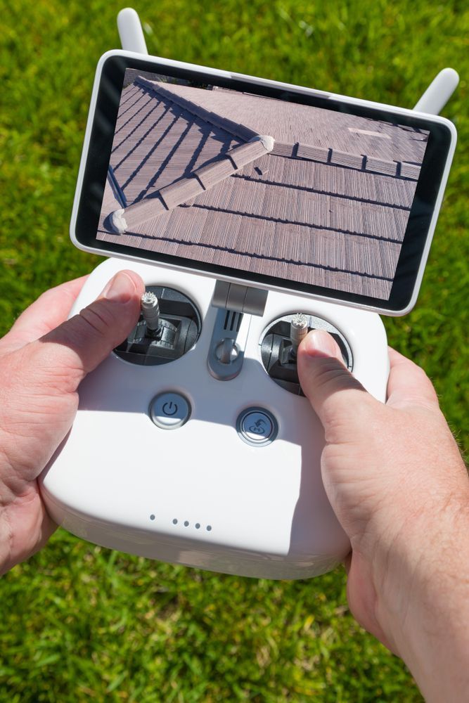 Hands holding a white drone controller with a phone showing a roof view, over green grass