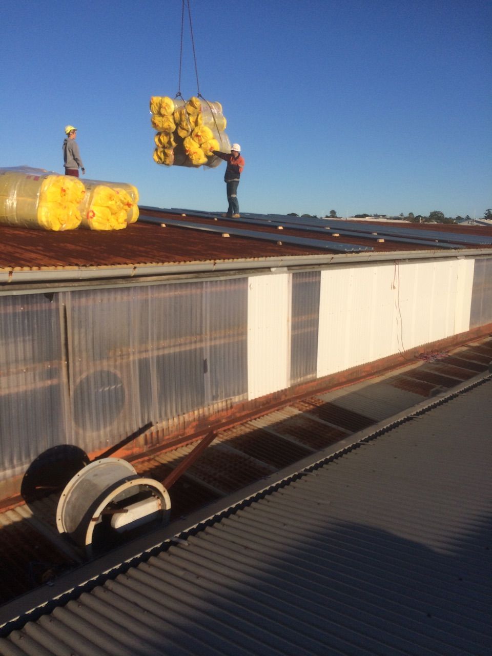 Two workers lifting yellow insulation on a rooftop under a clear blue sky