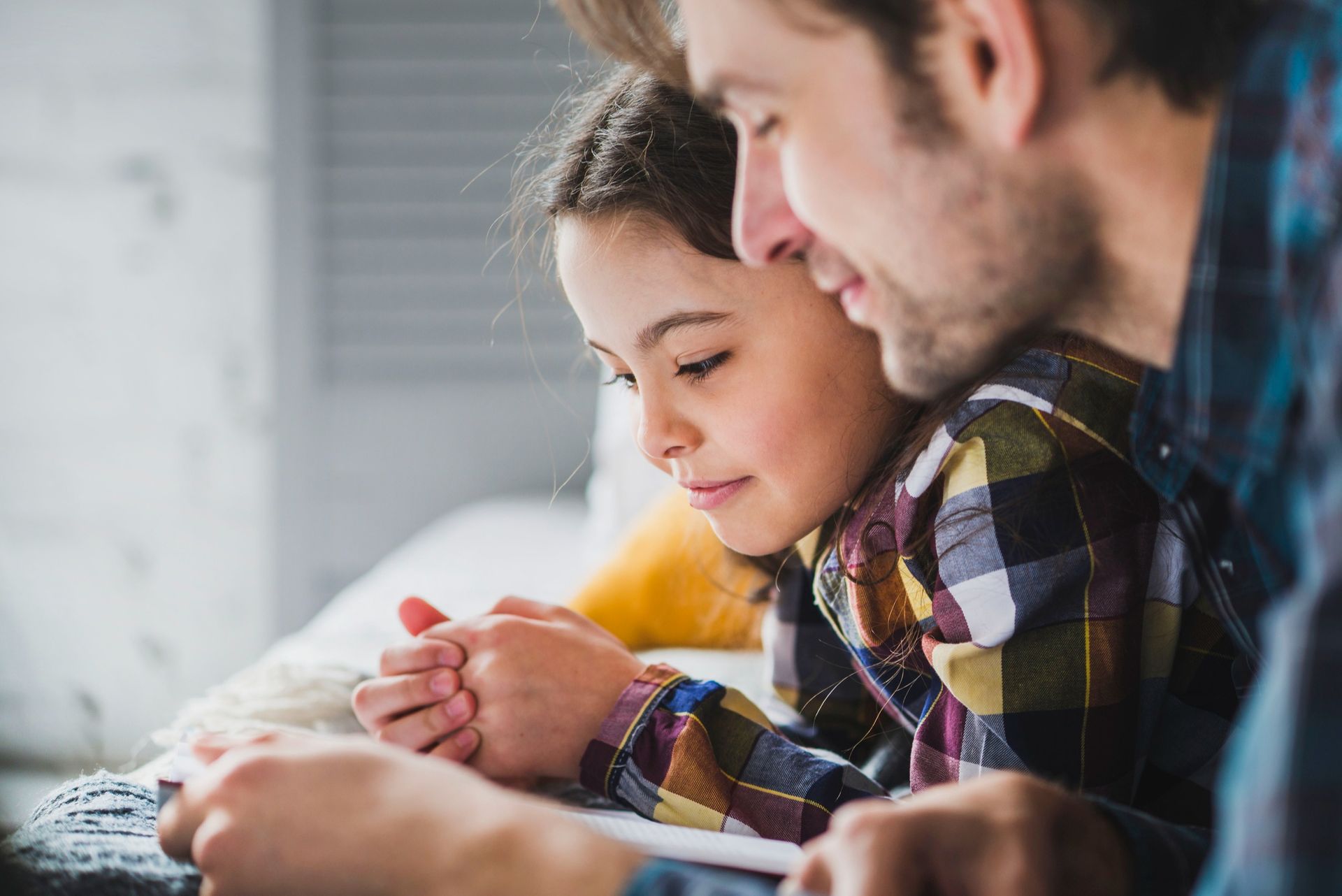 Man and child reading together, smiling. Girl's hands clasped, wearing plaid shirt. Indoors.