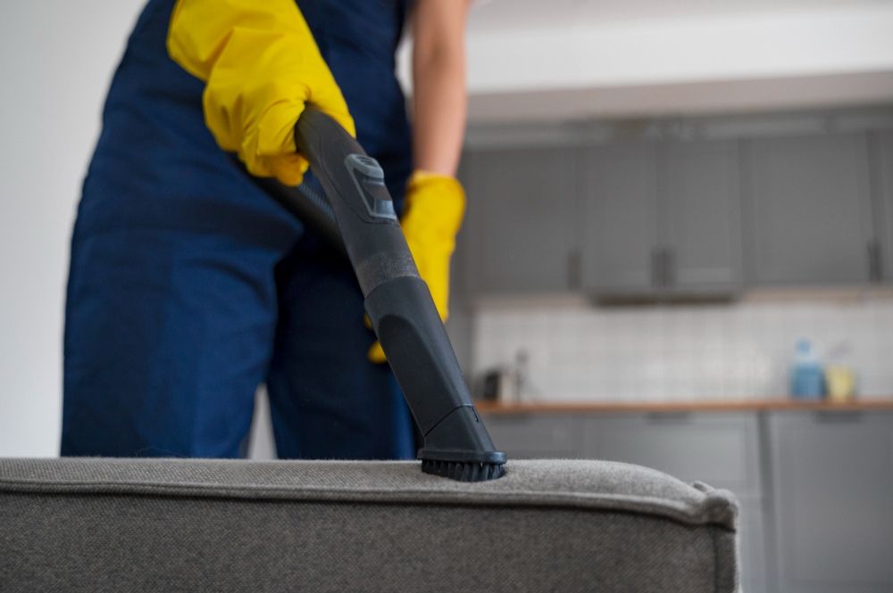 Person vacuuming a gray couch with a black vacuum attachment, wearing yellow gloves and a blue jumpsuit, indoors.