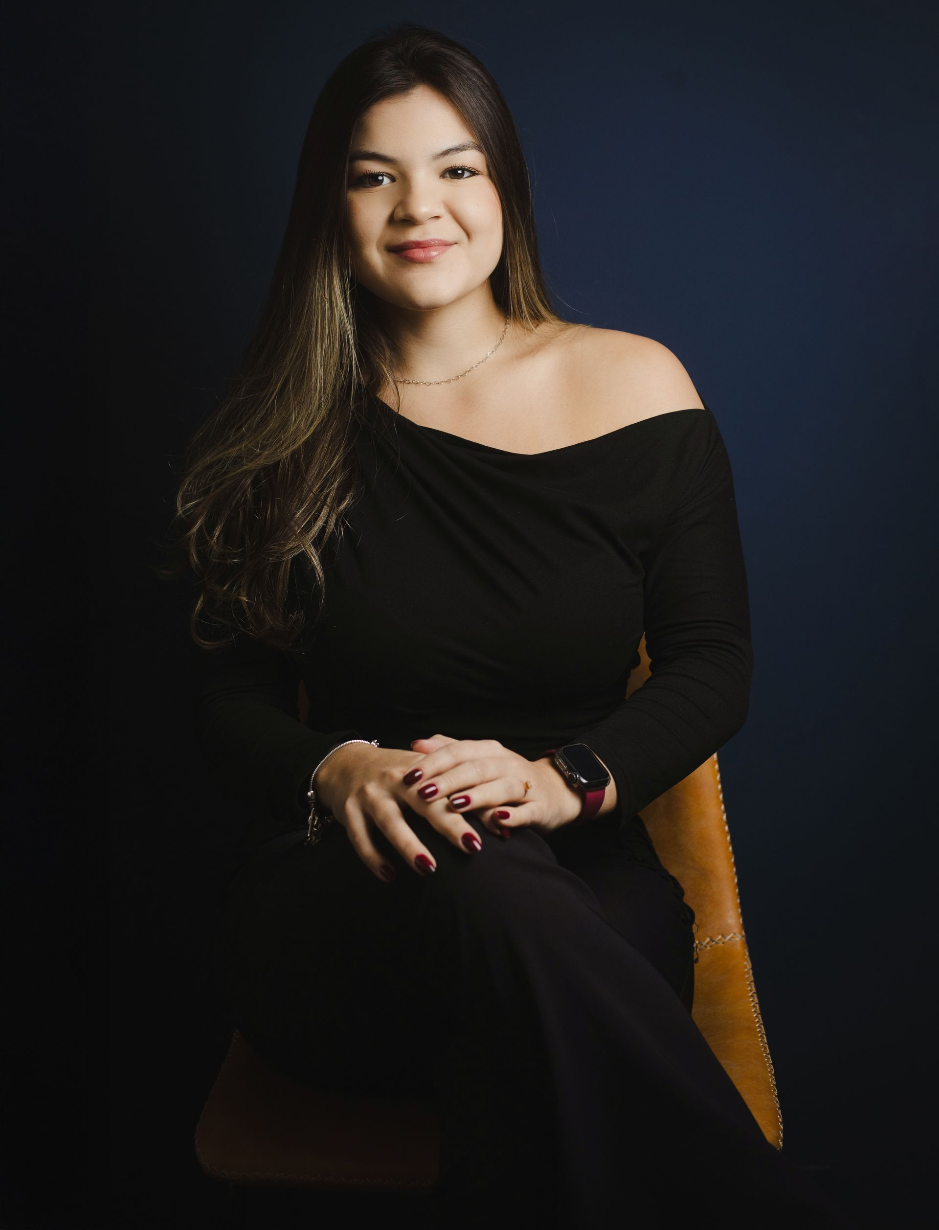 Woman in black off-shoulder top sits on chair, smiling. Dark blue background.