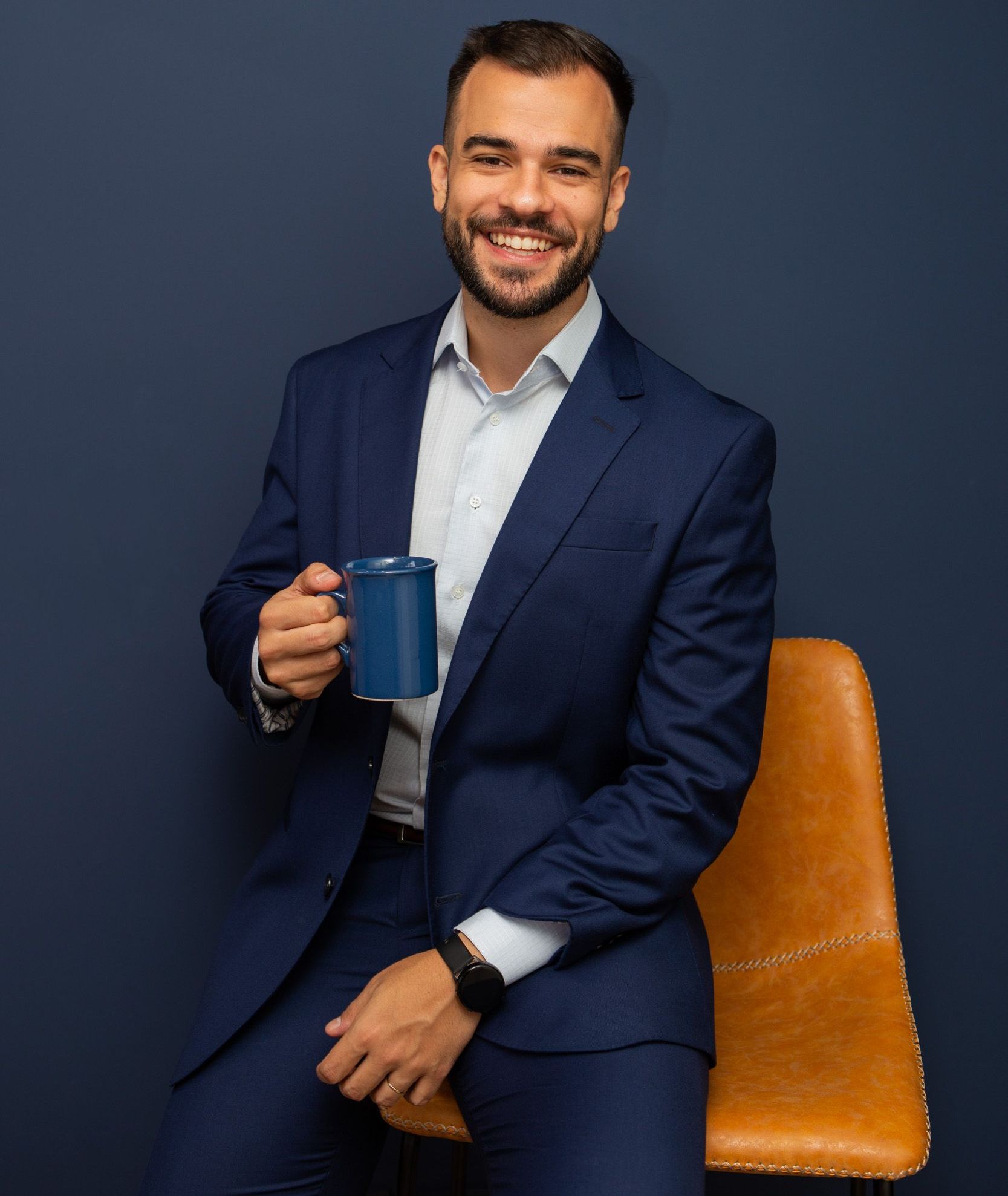 Man in blue suit, smiling, holding a blue mug, seated on a brown chair, against a dark blue wall.