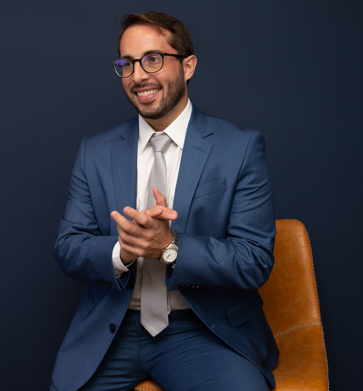 Man in a blue suit and glasses, smiling, clapping his hands. Sitting on a tan chair against a navy background.