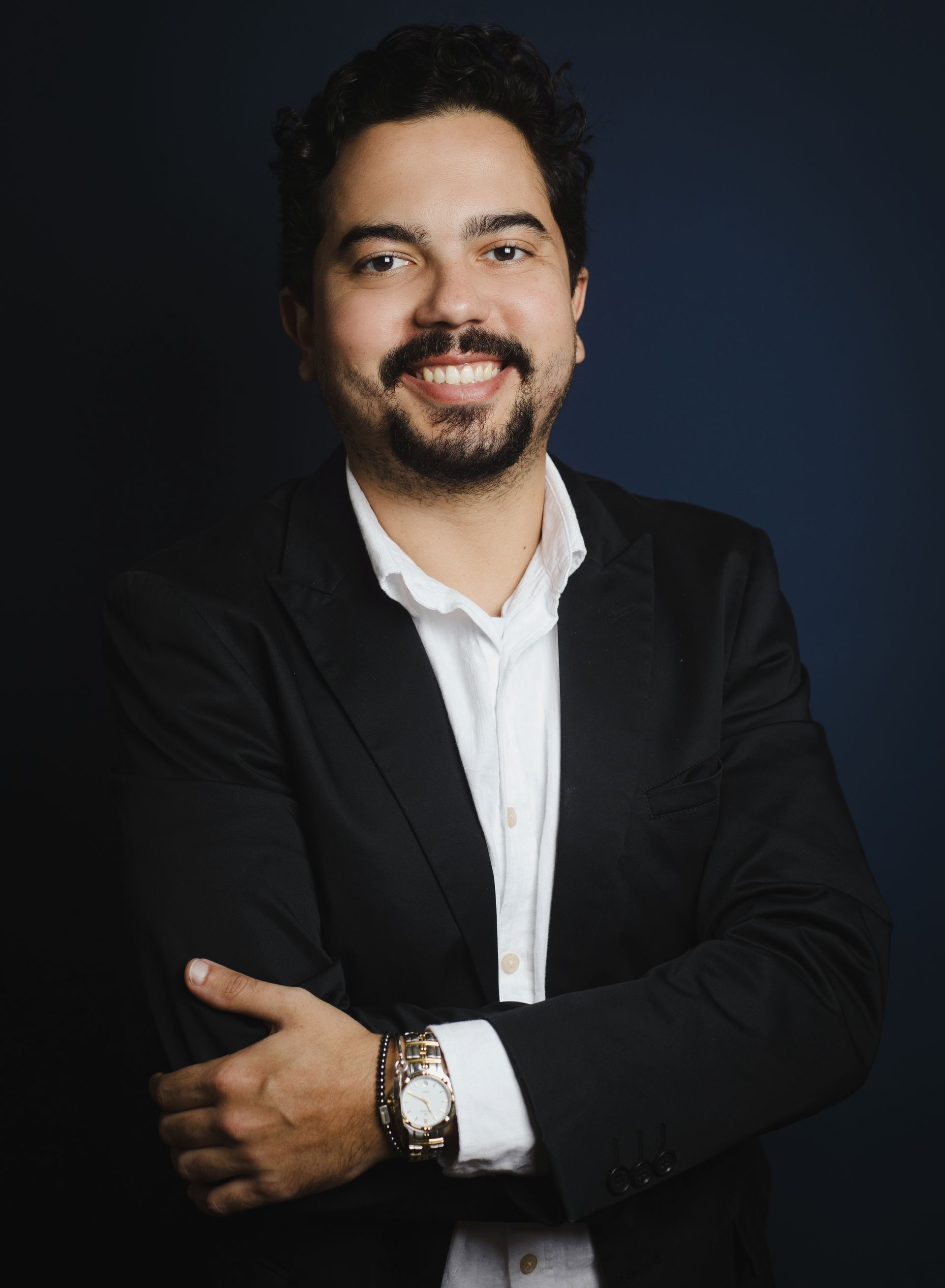 Man in black suit and white shirt smiles with arms crossed, watch on wrist, dark blue background.