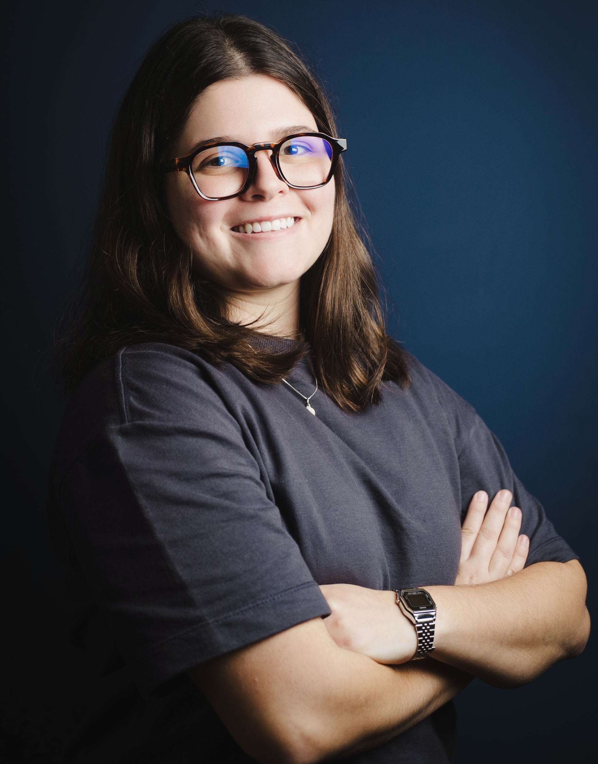 Woman with glasses, smiling with arms crossed, wearing a dark gray shirt against a dark blue background.