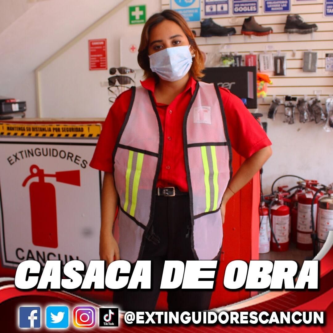 Una mujer con una mascarilla y un chaleco rosa se para frente a un extintor de incendios.