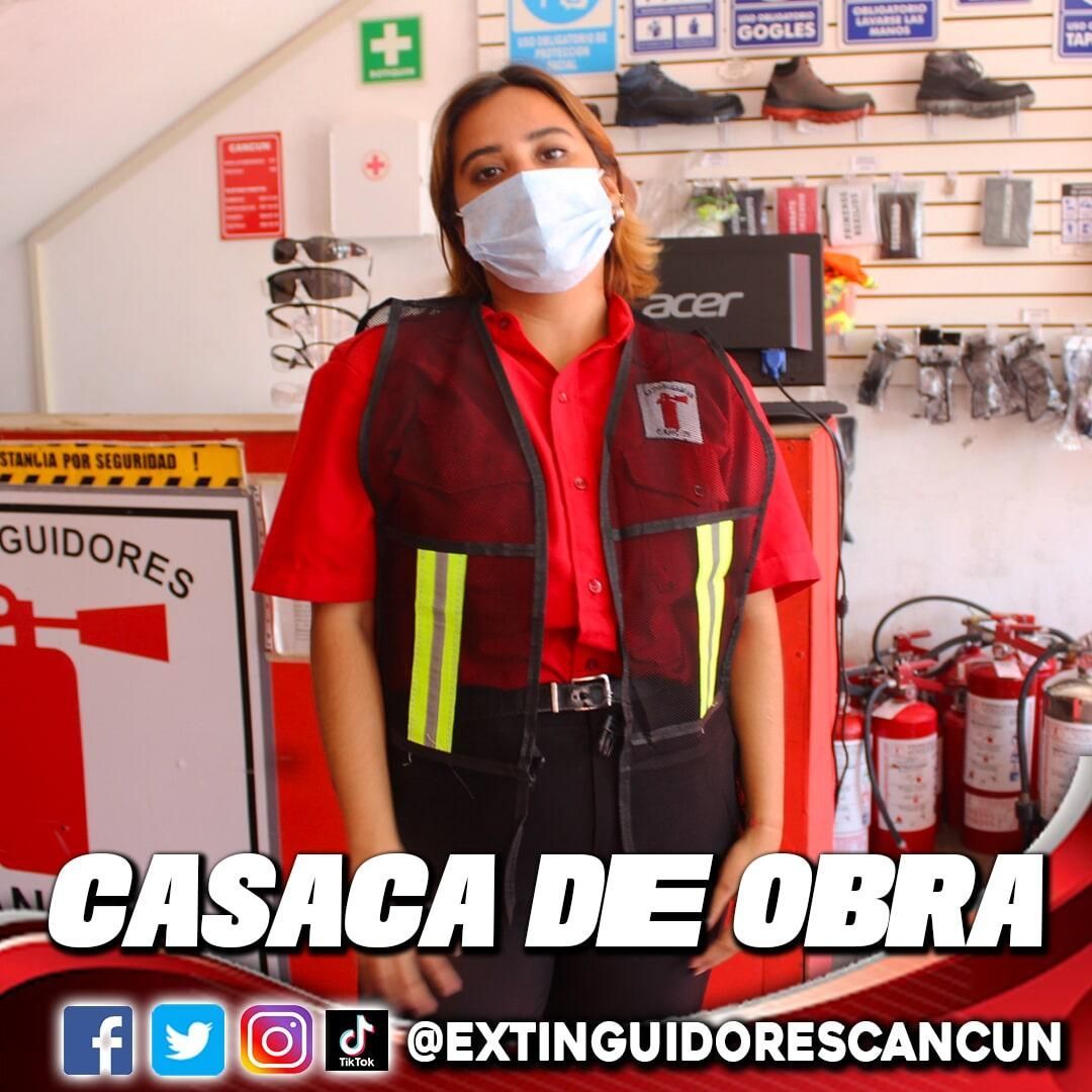 Una mujer con una máscara y una camisa roja está parada frente a los extintores.