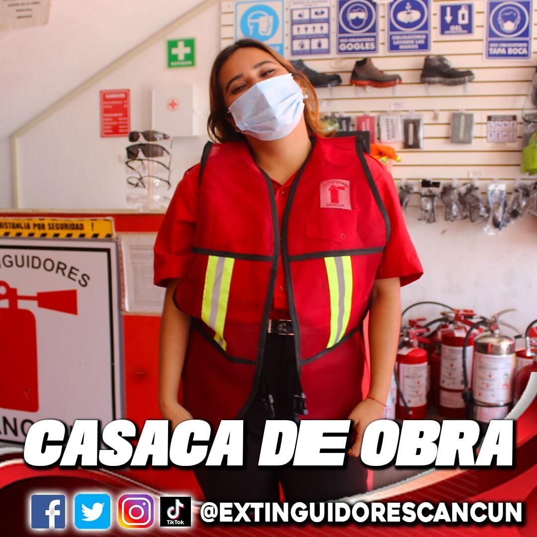 Una mujer con una máscara y un chaleco rojo se encuentra frente a los extintores.