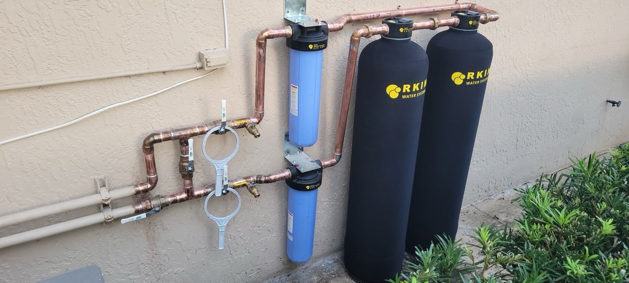 Outdoor water filtration system with copper pipes and three tall filter tanks against a house wall