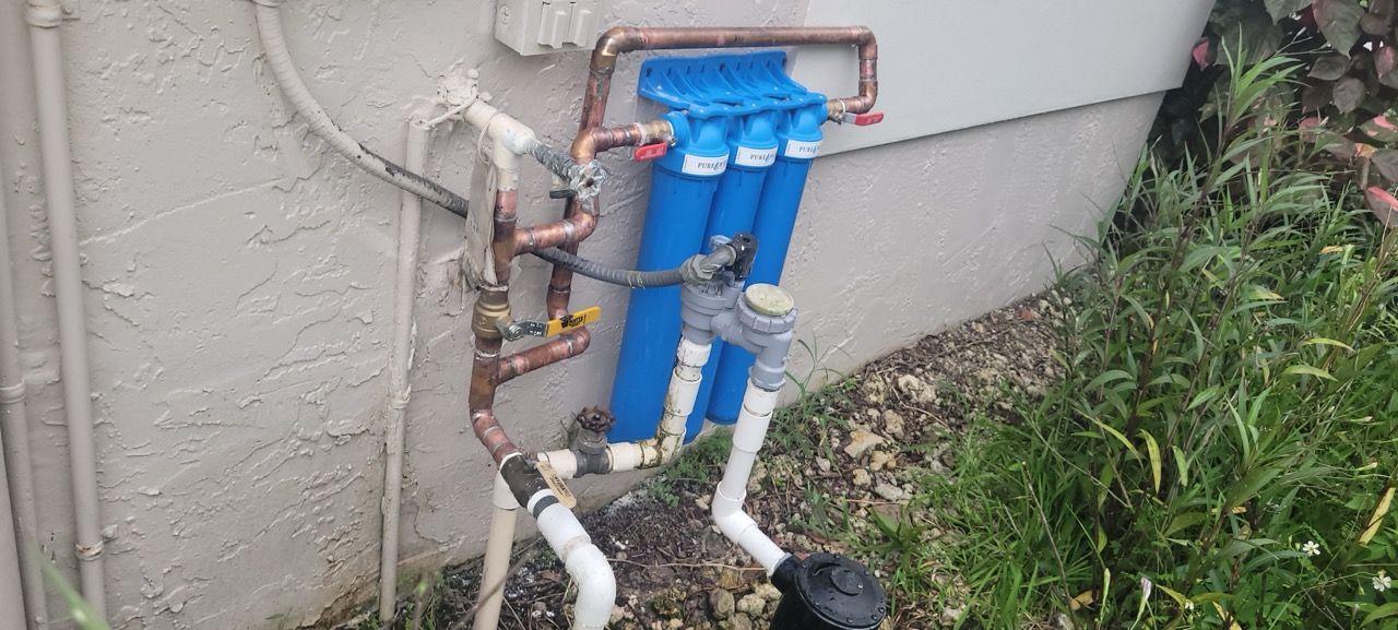 Blue water filtration system mounted on an exterior wall with copper pipes and white hoses