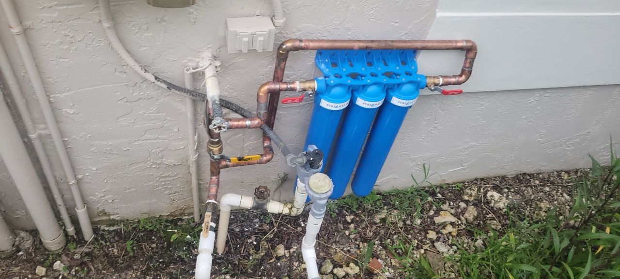 Blue water filter manifold and exposed pipes mounted on an exterior house wall