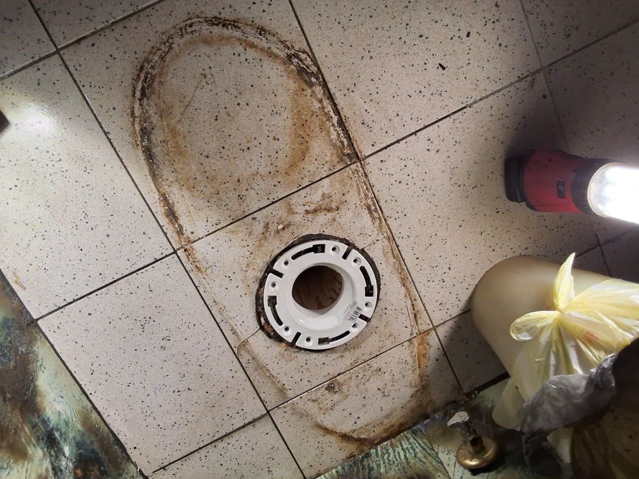 Bathroom floor with a drain and rust stain, plus a trash bag and pink shoe at the edge.