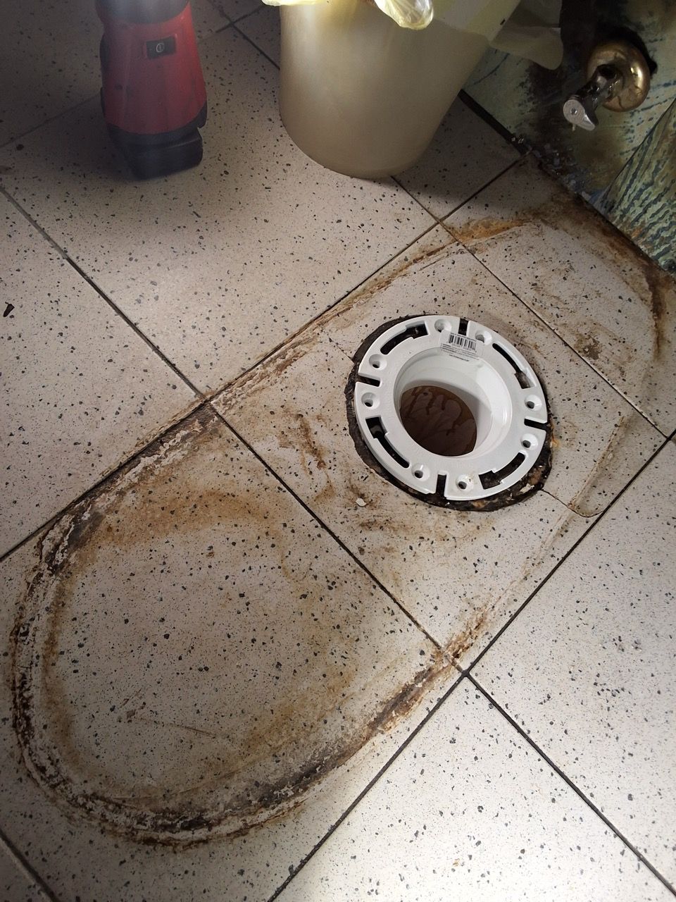 Dirty white toilet tank lid lying on speckled tile floor beside a toilet drain opening.