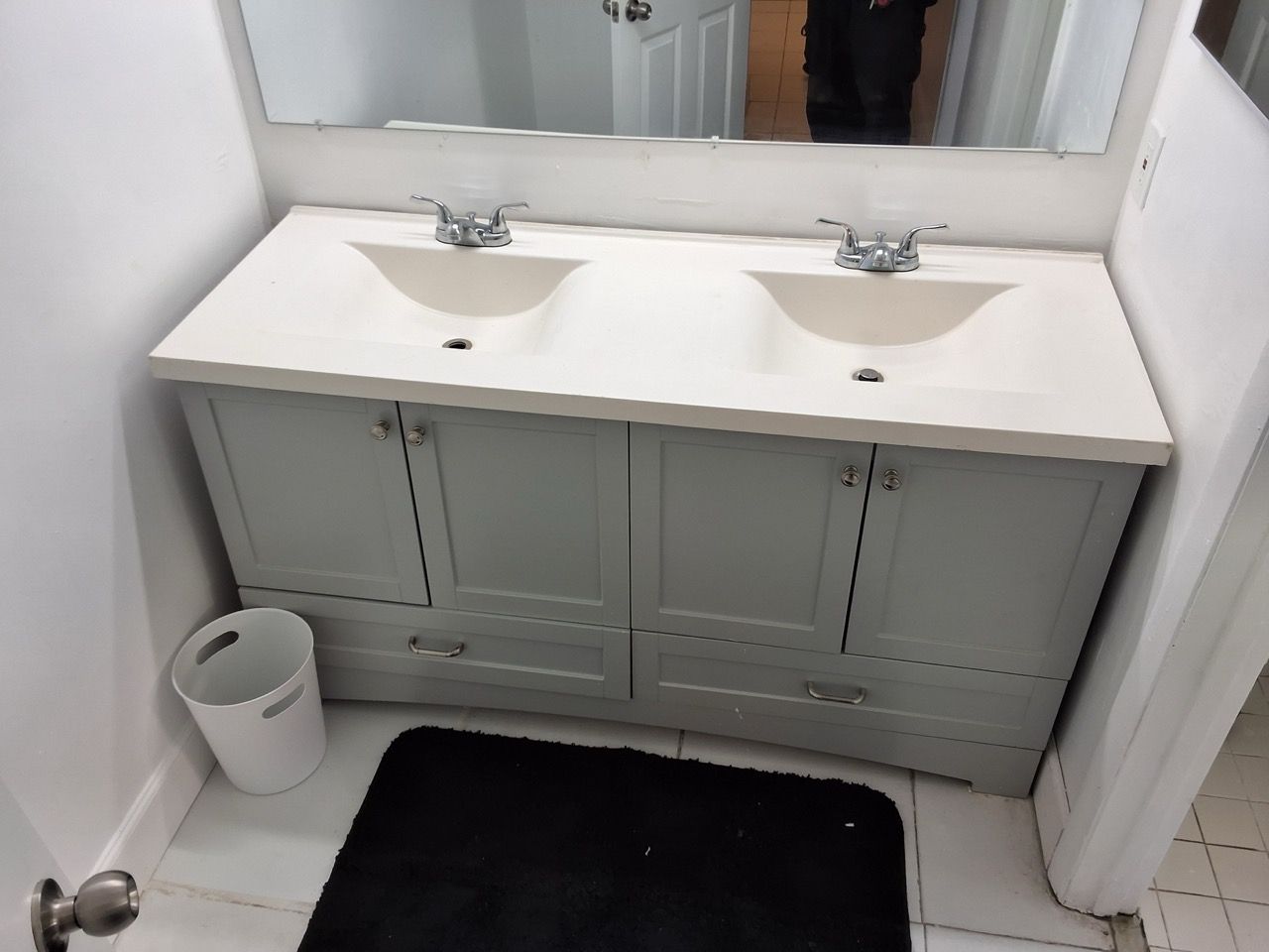 Small bathroom with double sink vanity, mirror, black bath mat, and a trash can.