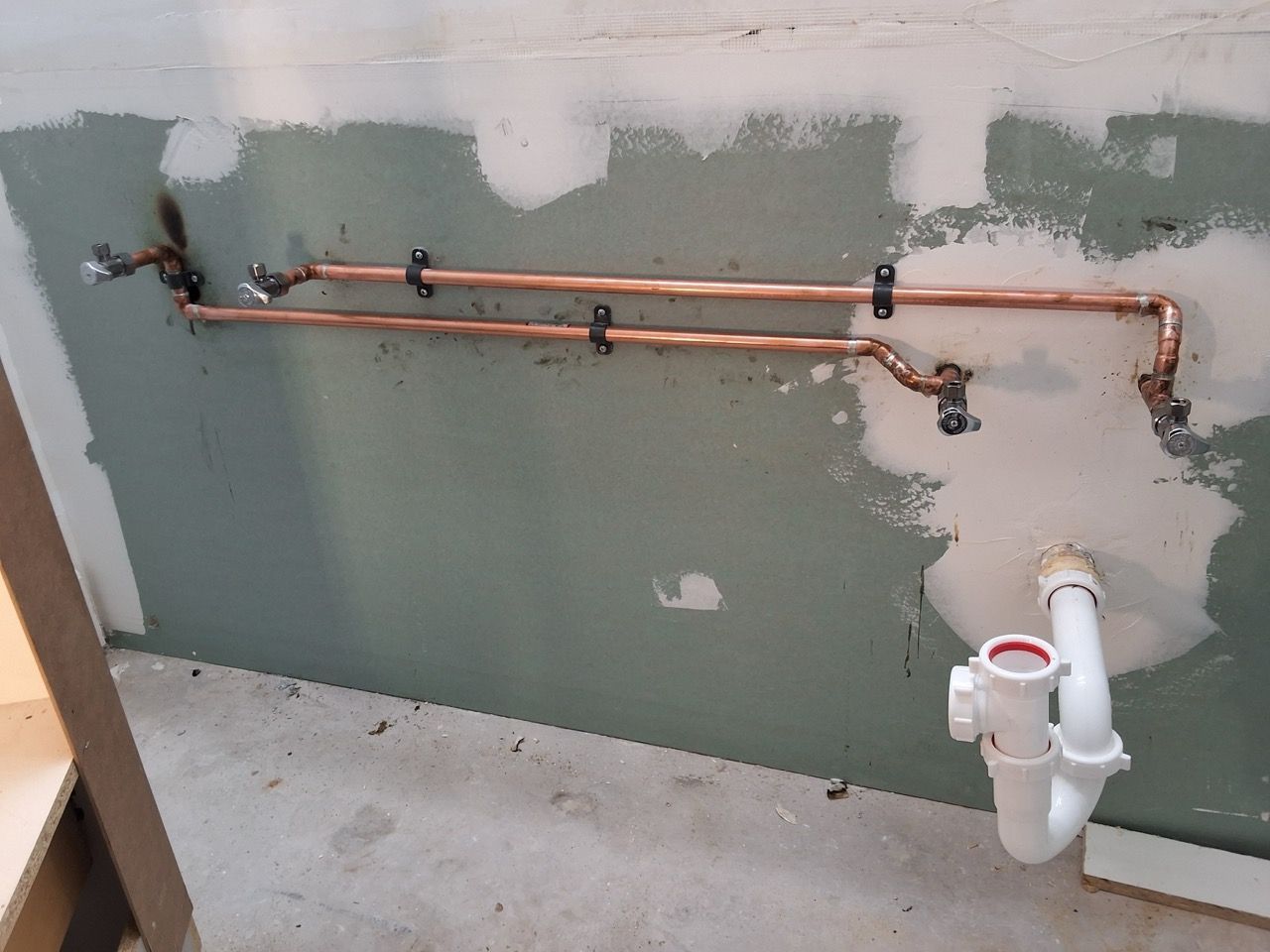 Copper plumbing pipes on a green wall above a concrete floor, with white PVC drain pipes below