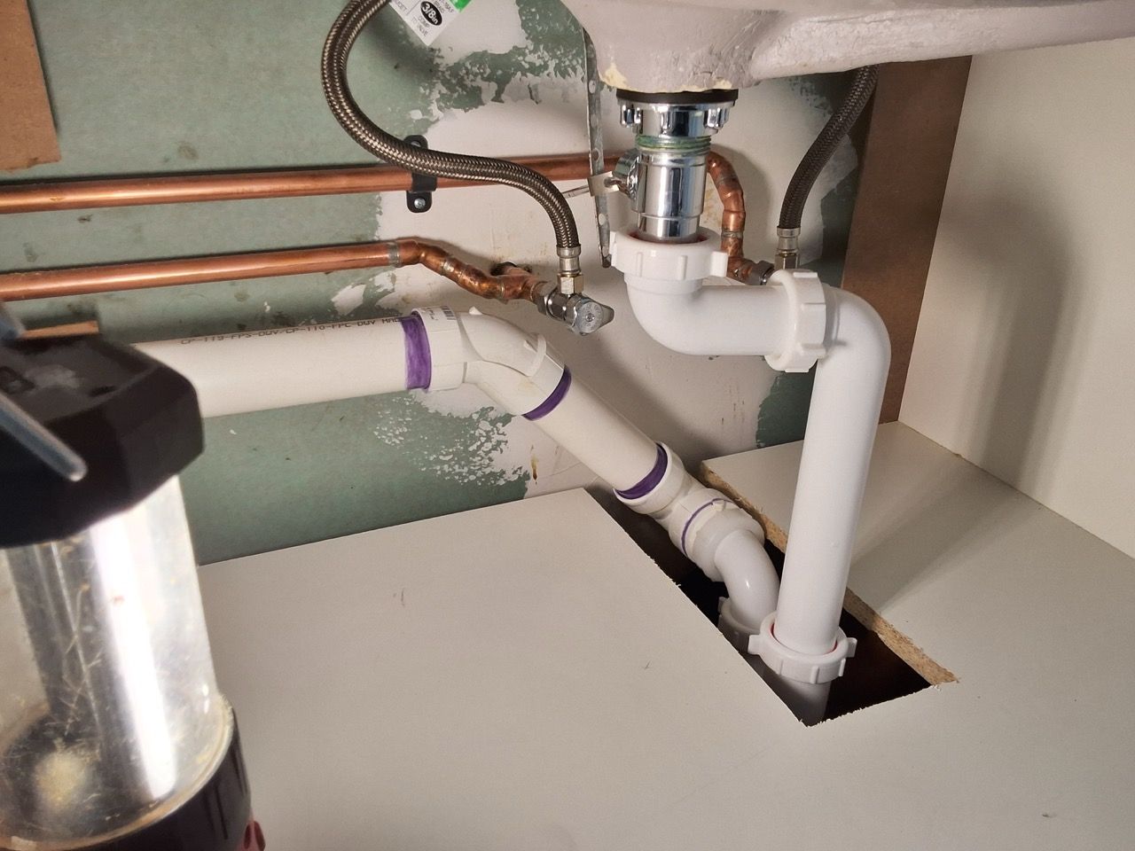 Under-sink plumbing with white PVC pipes, copper lines, and a cutout opening in the wall/floor.