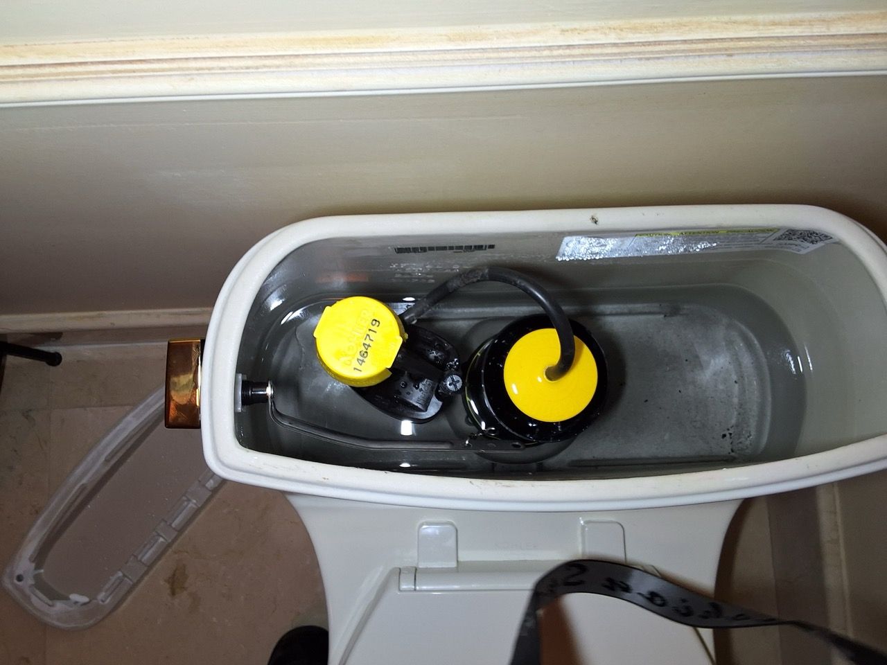 Open toilet tank with two black float valves and yellow caps inside.