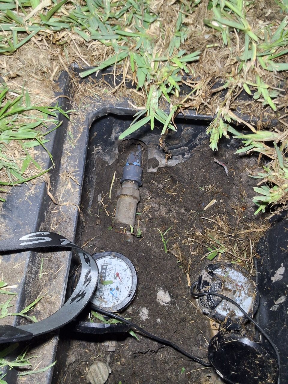 Damaged outdoor water meter box with exposed pipes, mud, and grass around the opening