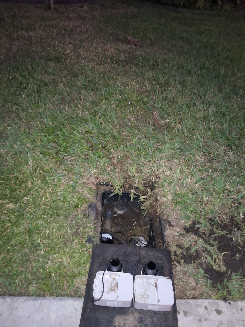 Open ground utility box in a grassy yard at night, with two white covers and exposed pipes inside.