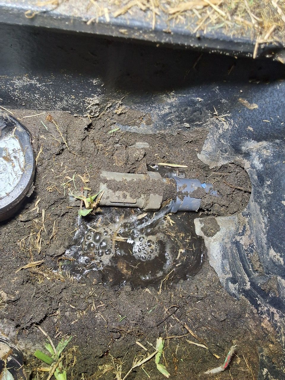 Dirty ditch drain with a small metal pipe and muddy water, surrounded by debris and grass.