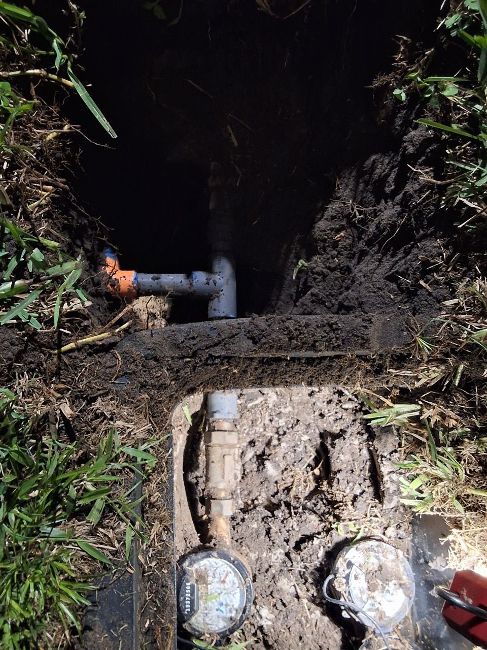 Close-up of a muddy utility pit with two round meter covers, a pipe, and vegetation around the opening