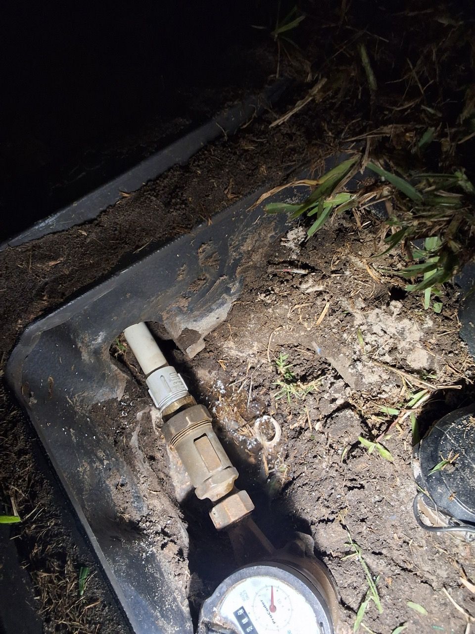 Broken pipe valve in a roadside drainage ditch at night, lit by a flashlight.