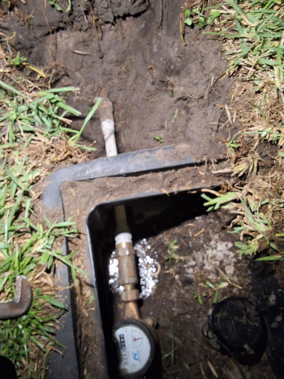 Open metal utility box in muddy ground with exposed pipe and valve amid grass