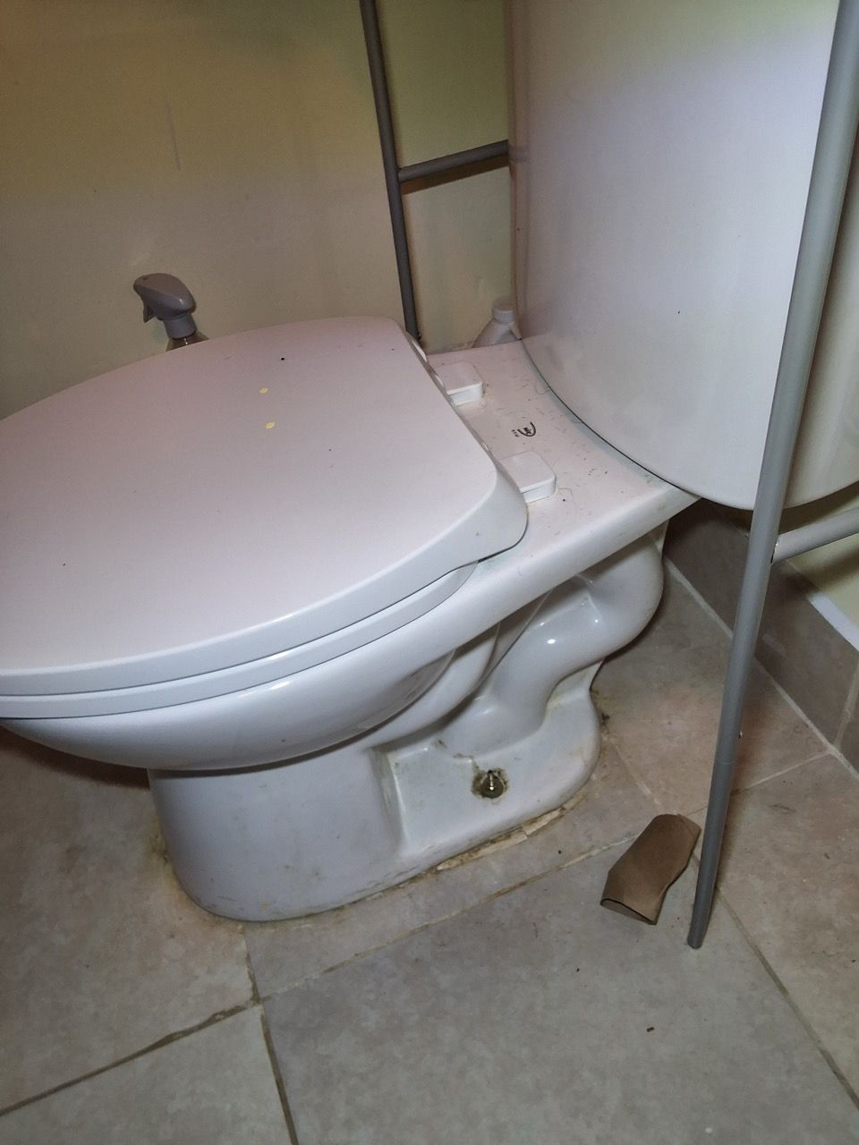 White toilet in a bathroom corner with a loose seat and visible damage near the base.