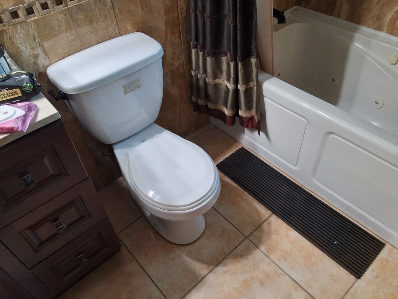 Bathroom with white toilet beside a tub, dark bath mat, and wooden vanity on tiled floor.
