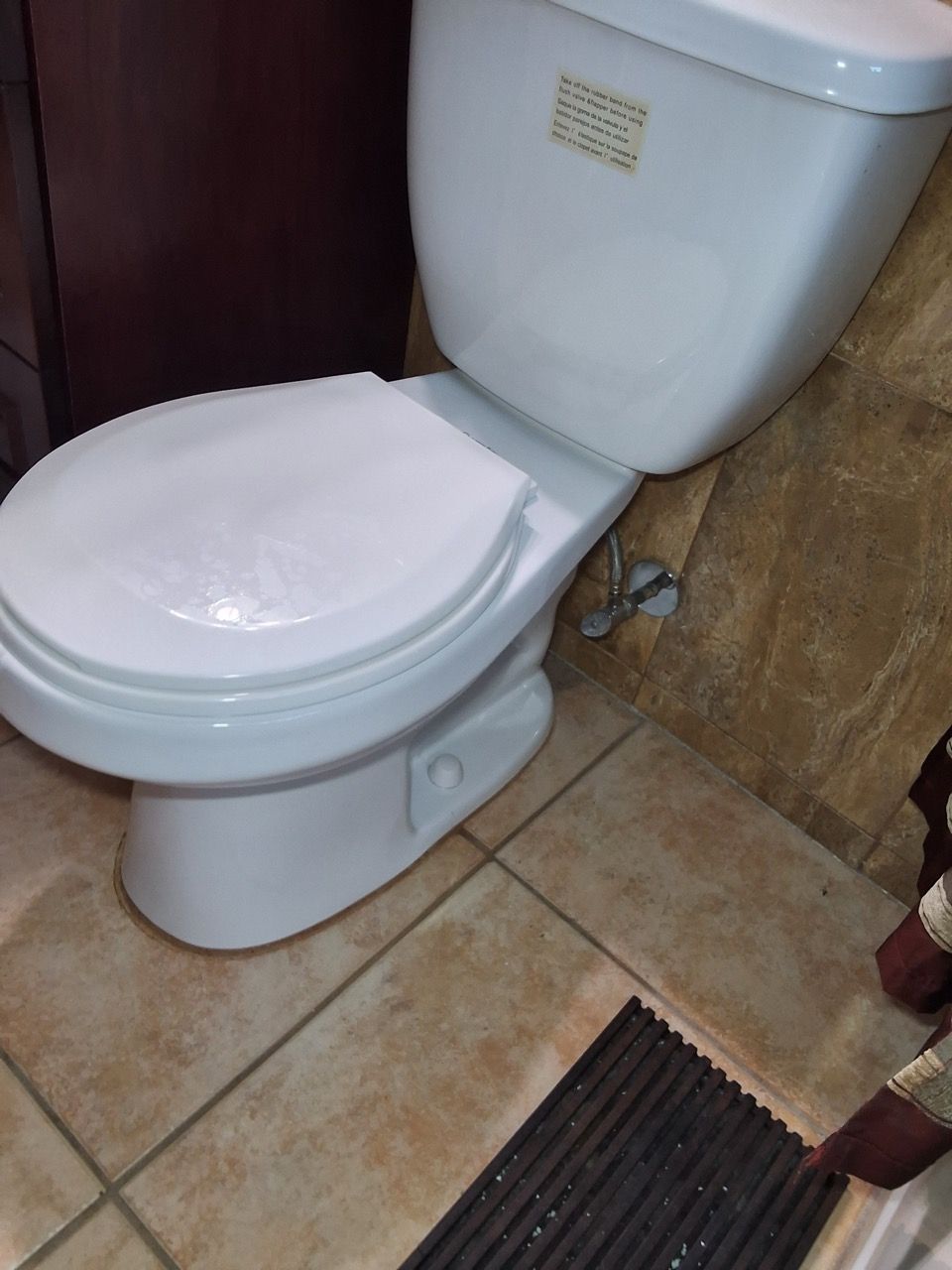 White toilet in a small bathroom with tan tile floor and a floor mat nearby