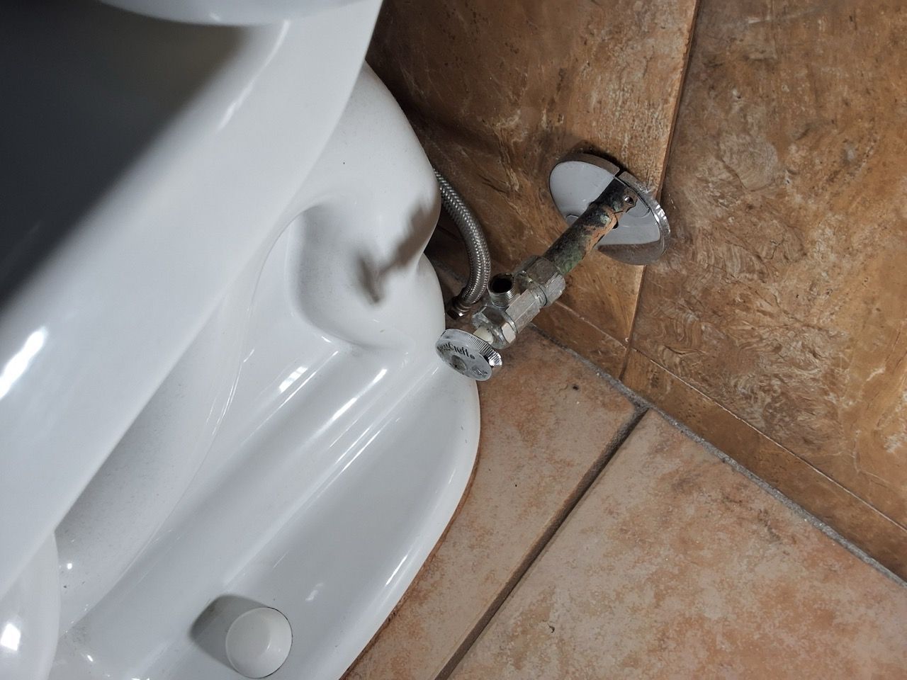 Close-up of a broken shower valve and pipe behind a toilet, with exposed plumbing and tiles.