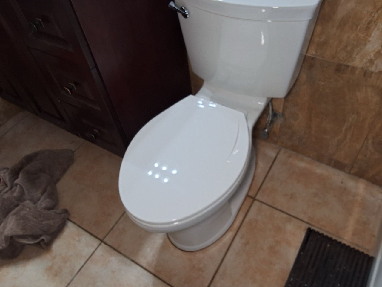 White toilet with closed lid in a tiled bathroom corner, beside a dark cabinet and floor vent.