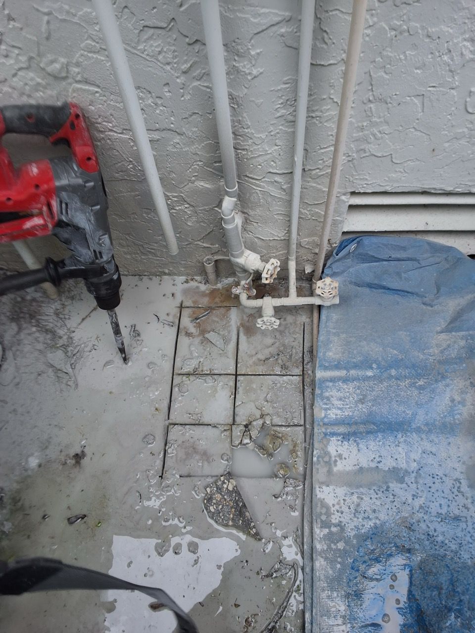 Broken white pipes beside a gray wall, wet tiled floor, and a blue tarp in a work area