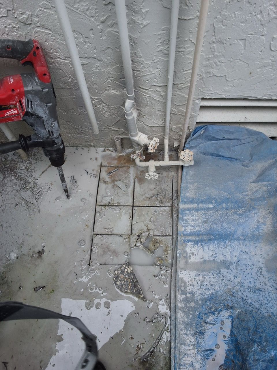 Damaged concrete floor with exposed pipes, debris, and a red tool on the left beside a blue tarp