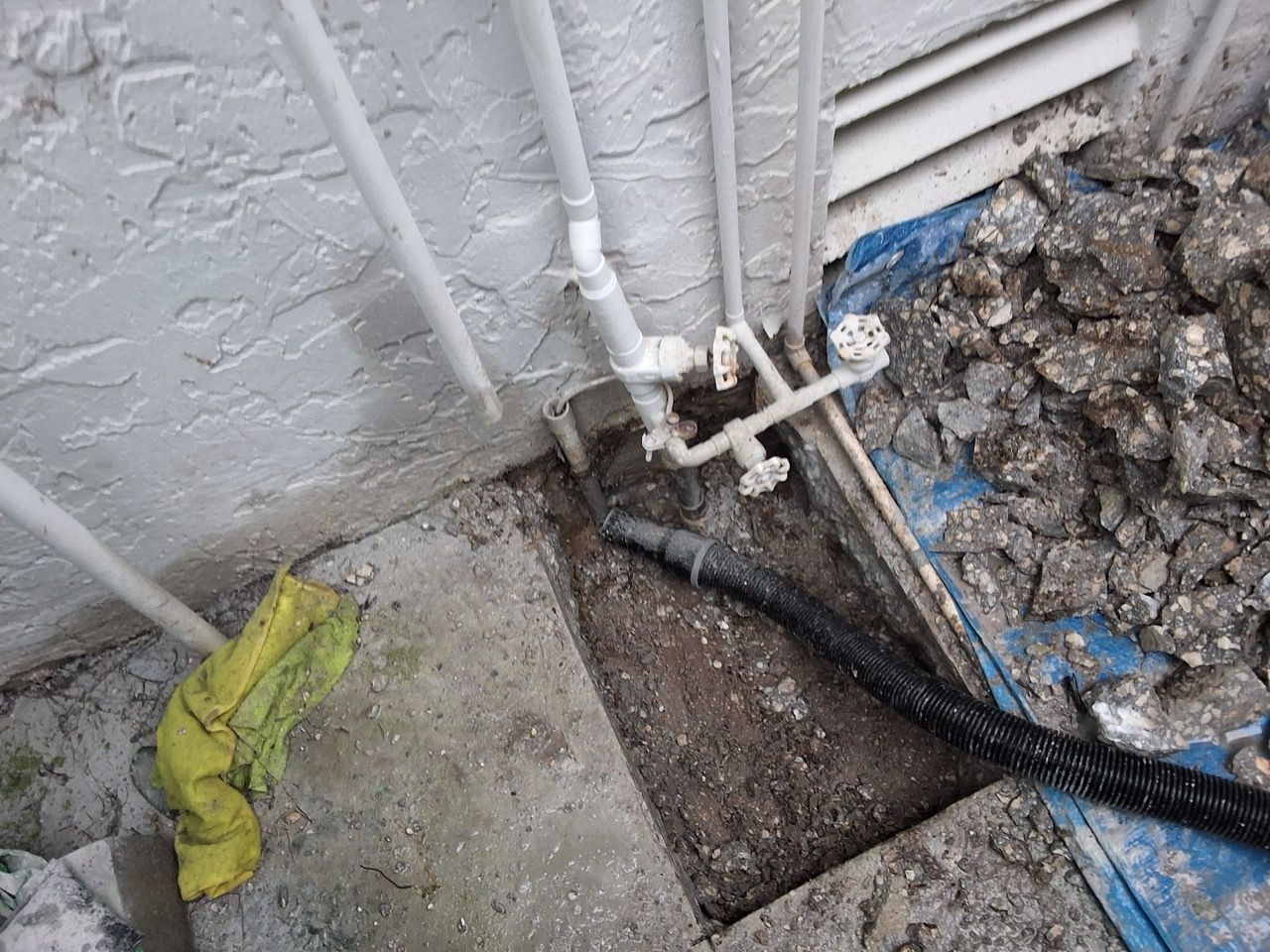 Broken outdoor pipe and exposed drain by a concrete wall, with a yellow cloth nearby.