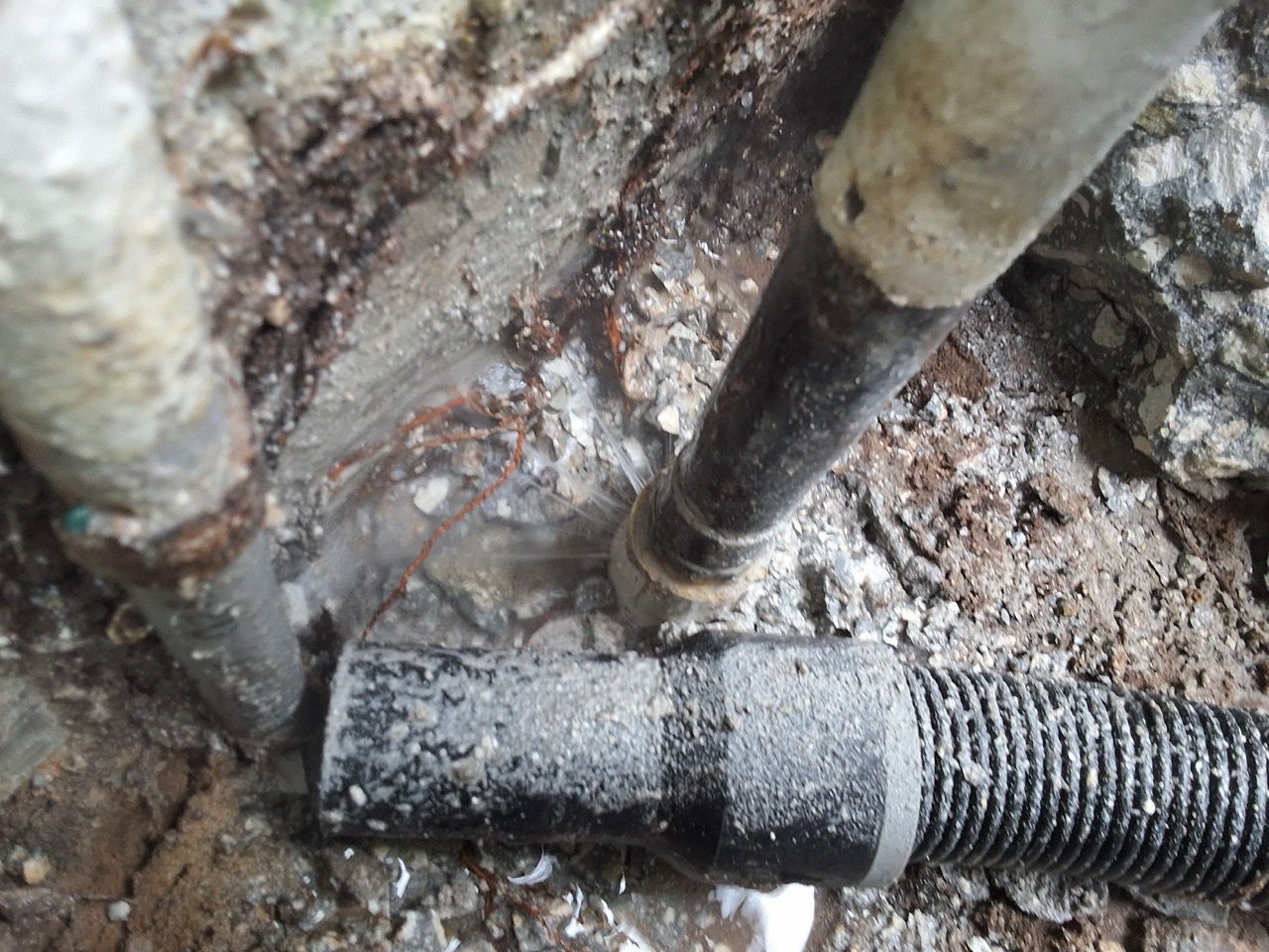 Damaged pipe joint in muddy ground with exposed black corrugated tubing and rusted metal pipes