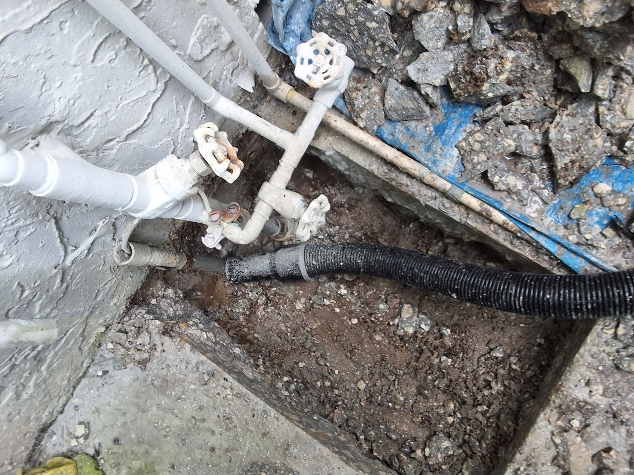 White pipes and valves beside a concrete wall, with a black corrugated cable in a dirt trench.