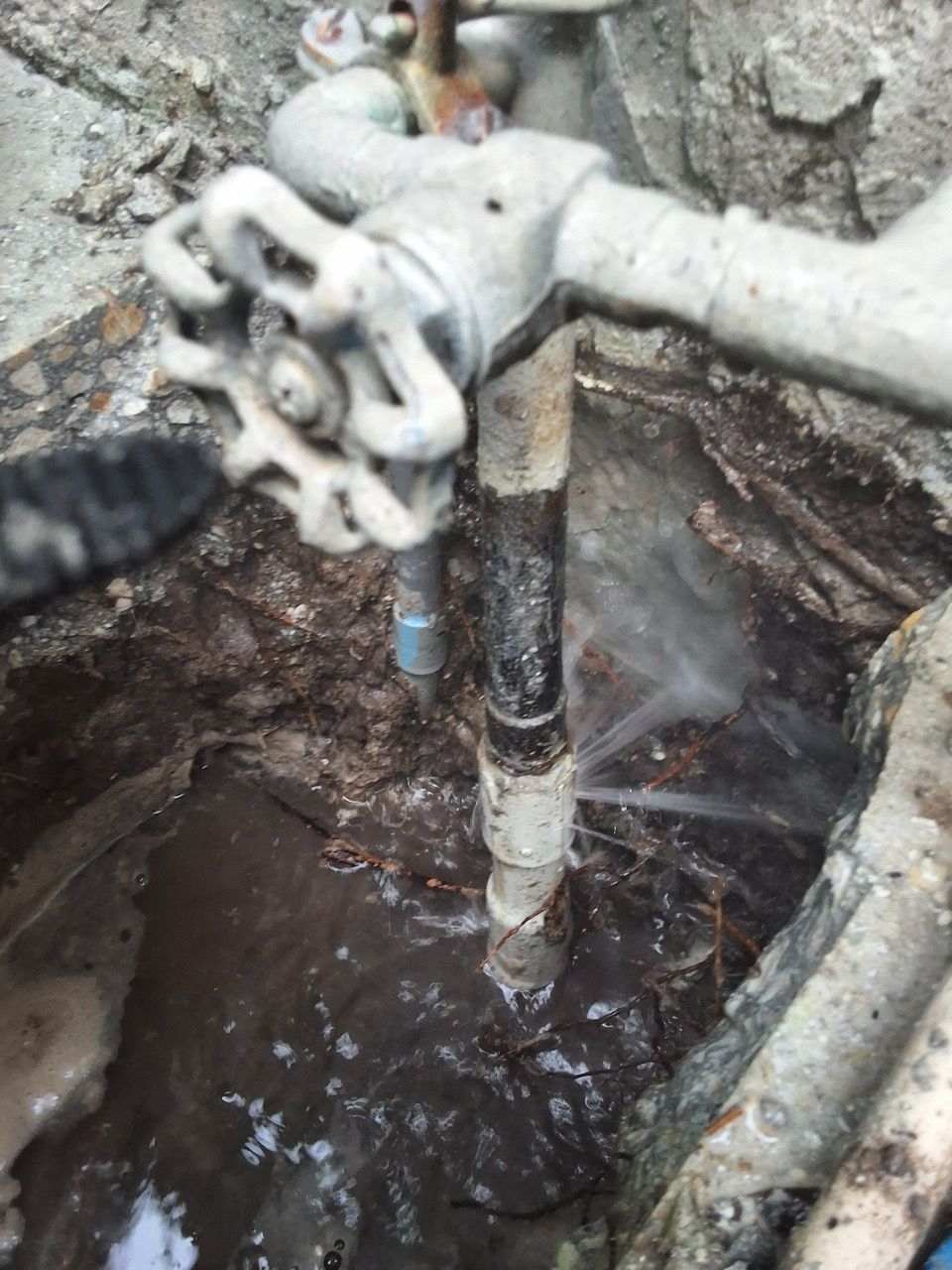 Plumbing valve and pipes in a muddy excavation hole, with water pooled around the pipe base