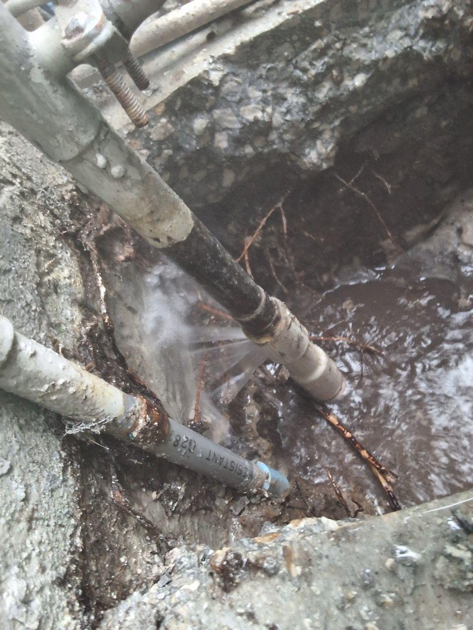 Water spraying from pipes into a muddy excavation pit with exposed metal pipes and concrete walls