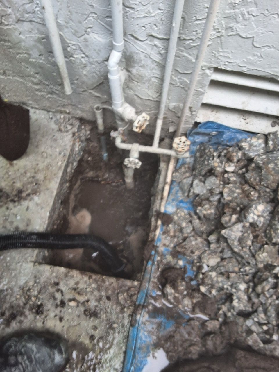 Damaged outdoor plumbing and a muddy excavation beside a wall, with exposed pipes and rubble.