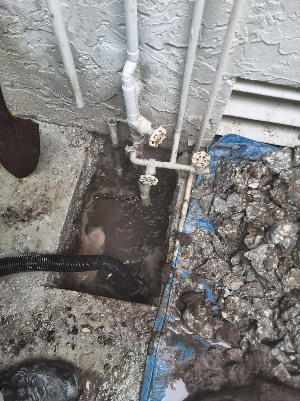 Open utility trench with exposed pipes, a valve, and muddy soil beside a concrete wall.