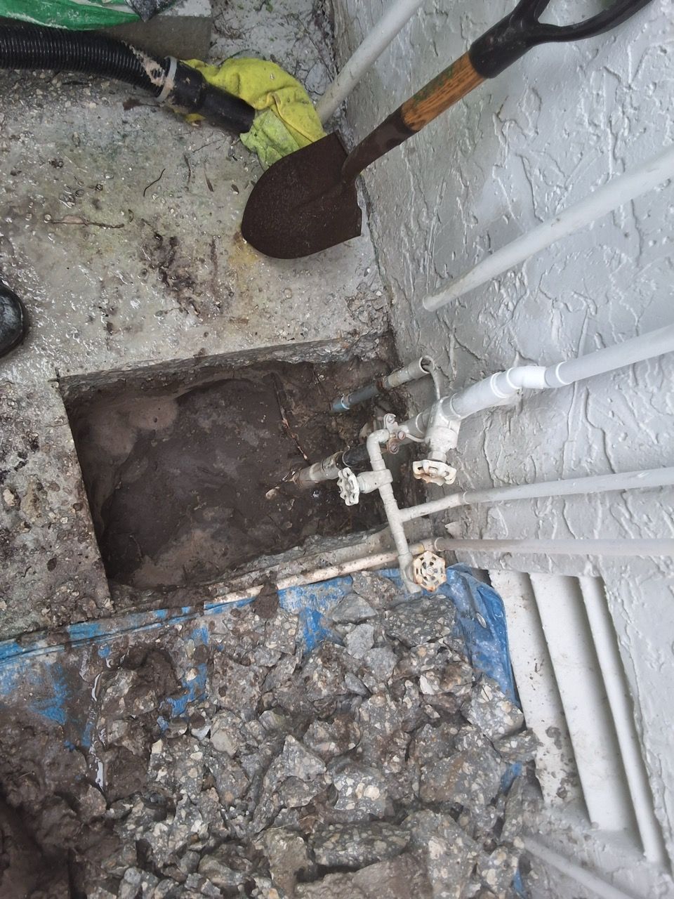 Exposed utility trench beside wall with pipes, valve, and a shovel on gravel debris