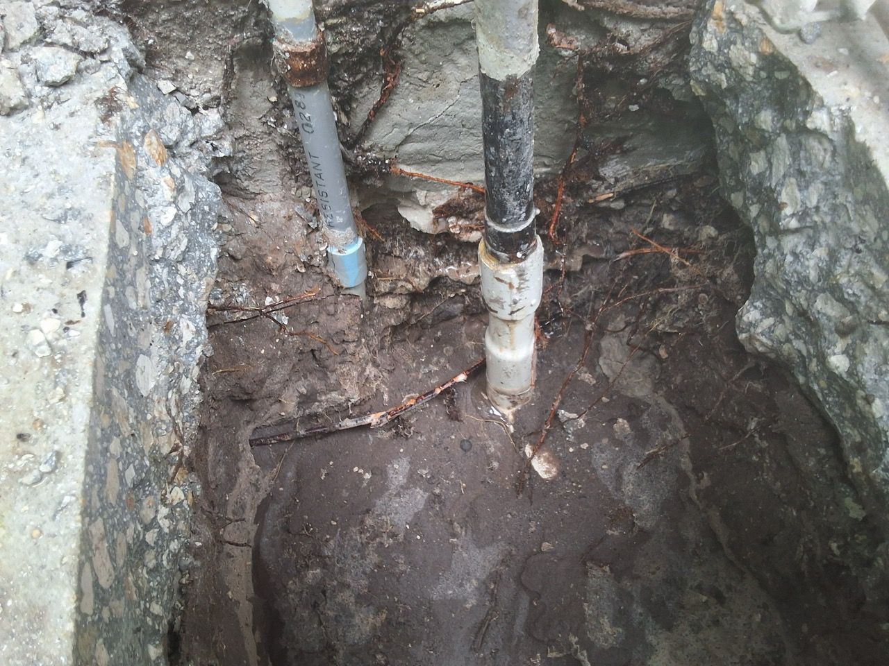 Open excavation with exposed underground pipes and muddy soil between stone walls