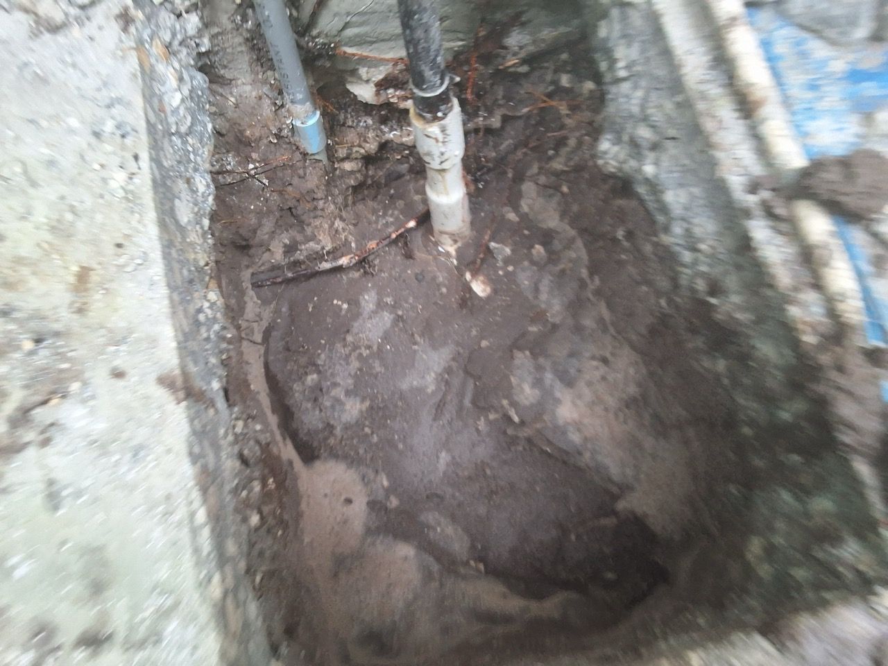 Dirty excavation pit with exposed pipes and muddy ground beside a wall